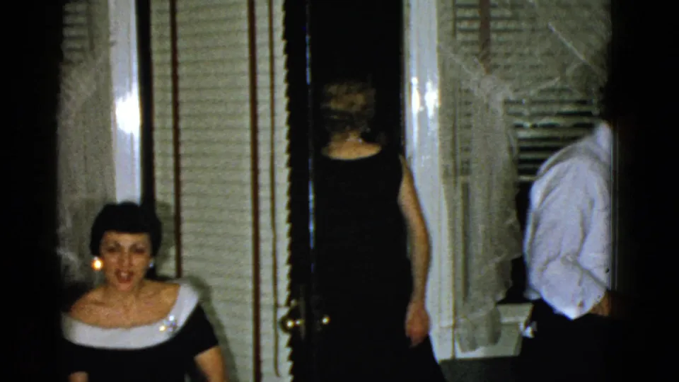 1951 Boston home-movie frame with a seated woman facing the camera as formally dressed adults move through the room.