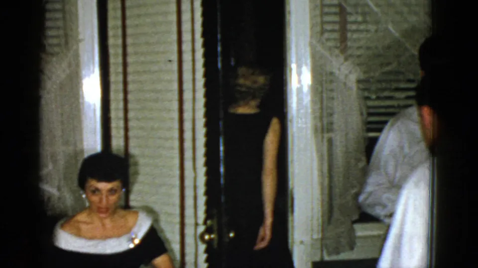 1951 Boston home-movie frame showing a seated woman and adults moving through a doorway in a formal home interior.