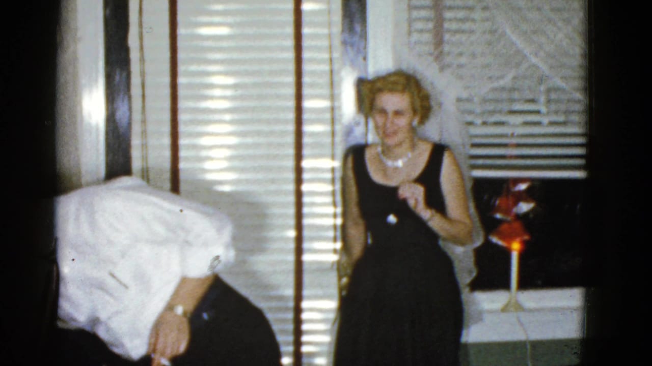 Adults at an indoor reunion, including a woman in a dark dress and a partially visible man bending near venetian blinds.