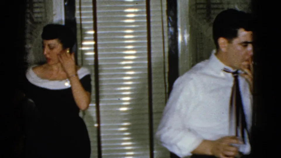A woman in a dark dress with a light neckline stands near venetian blinds in the 1951 Boston reunion footage.