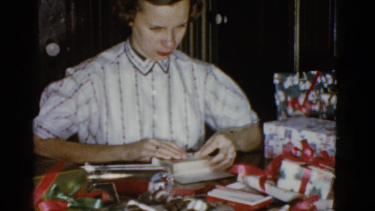 The woman folds wrapping paper around a small box while red-ribboned gifts sit on the table in the dim interior.