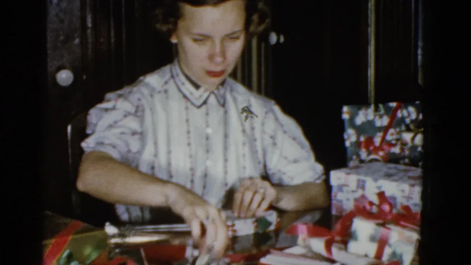The woman reaches toward supplies on the tabletop, surrounded by holiday wrapping paper, ribbon, and already wrapped gifts.