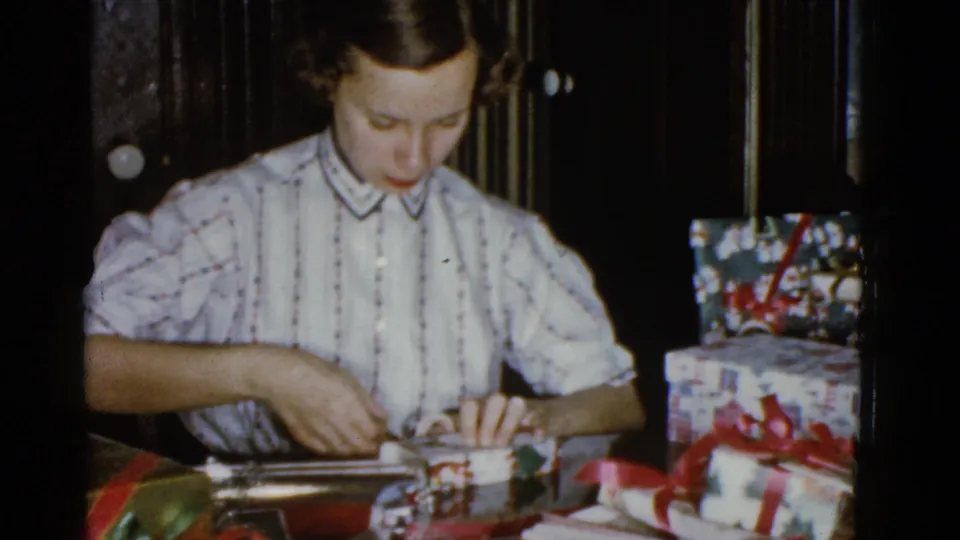 The woman looks down and works with her hands to secure a small wrapped gift, with Christmas packages visible to one side.