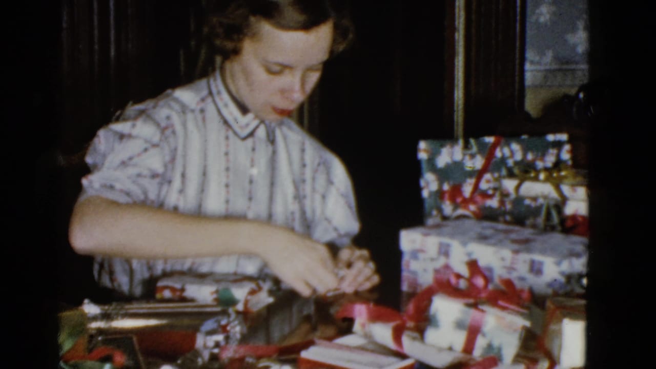 In 1951 Boston home-movie footage, a young woman in a striped blouse wraps a gift at a table with stacked presents nearby.