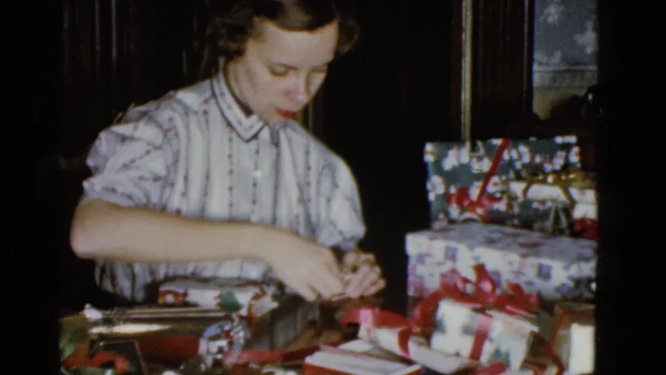 In 1951 Boston home-movie footage, a young woman in a striped blouse wraps a gift at a table with stacked presents nearby.