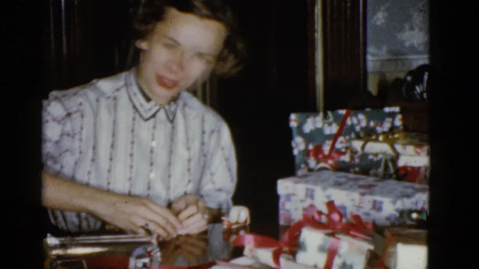 A young woman smiles during Christmas gift wrapping at a table, with decorated presents and red ribbons visible beside her.