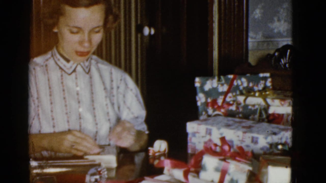 In 1951 Boston home-movie footage, a young woman in a striped blouse wraps a gift at a table beside red ribbon and wrapped boxes.
