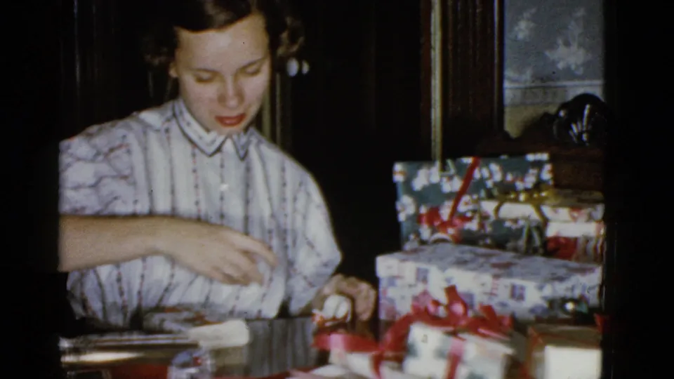 The woman continues wrapping gifts indoors, reaching across the table near red ribbon, paper, and wrapped Christmas boxes.