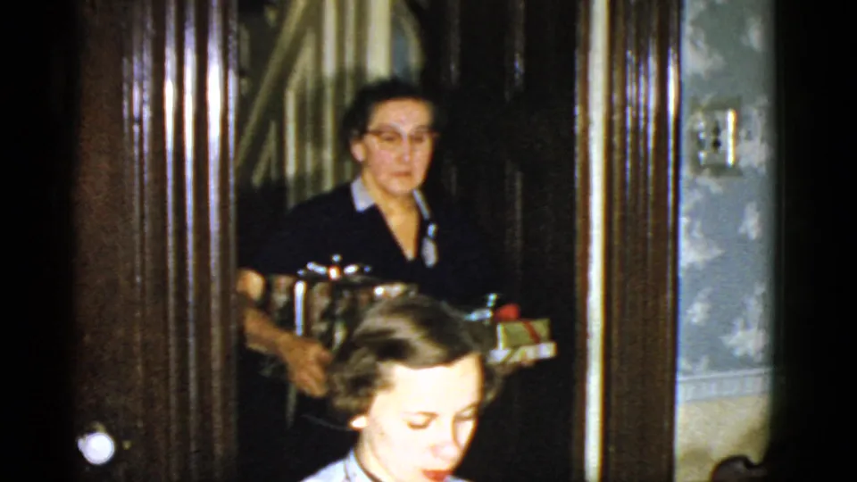 An older woman in glasses carries gifts through a doorway behind a seated person, with wood trim and patterned wall covering visible.