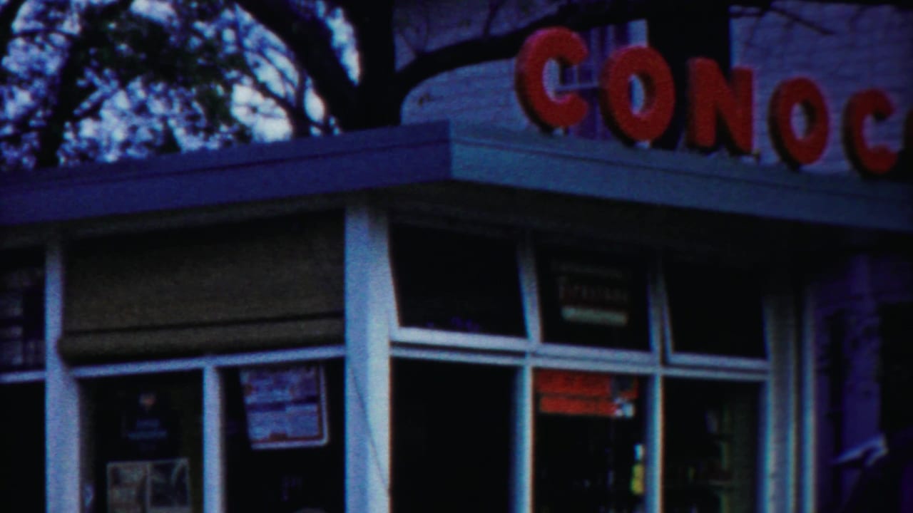 1958 roadside station view with red Conoco lettering, glass windows, white columns, and dark tree branches overhead.