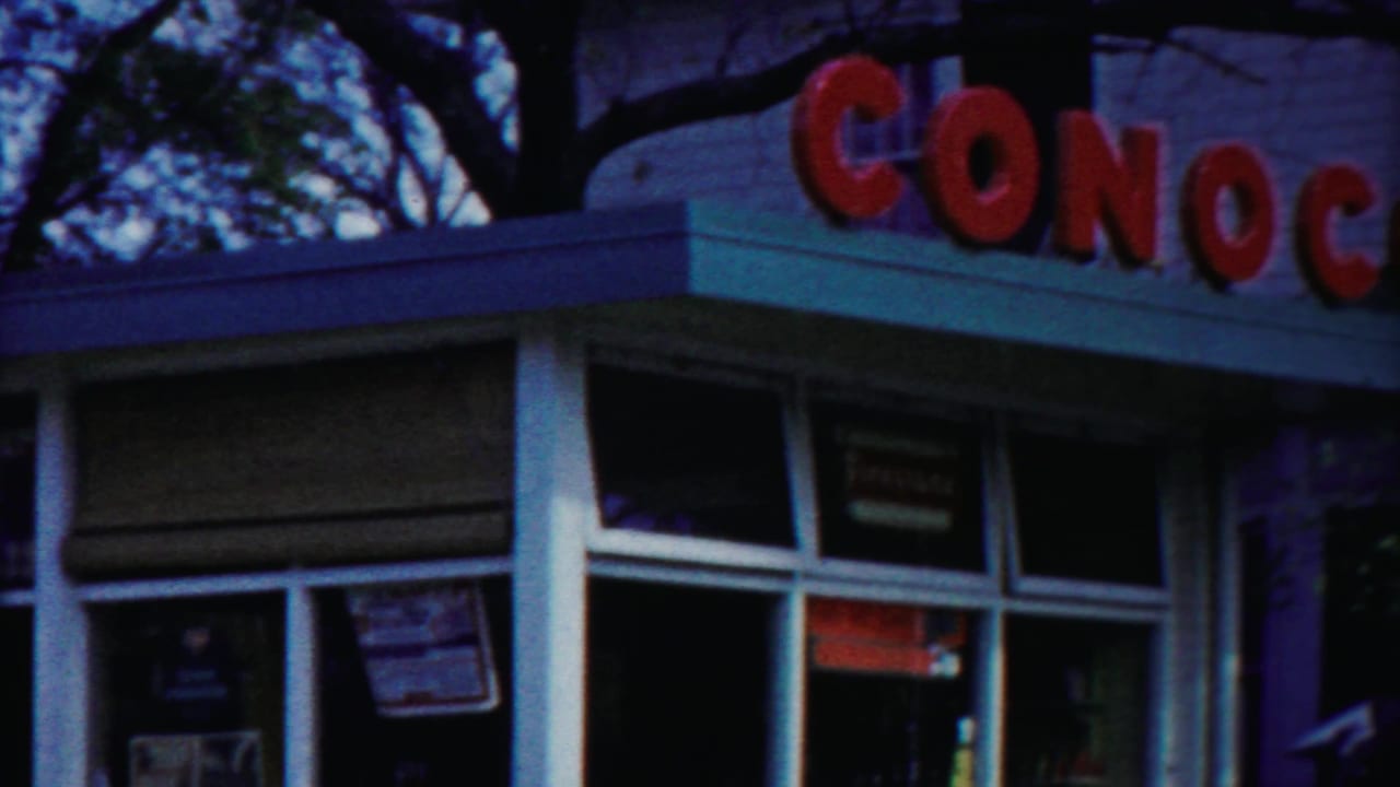 Angled 1958 view of the Conoco station corner, showing red exterior lettering, large glass panes, window placards, and the roof edge.