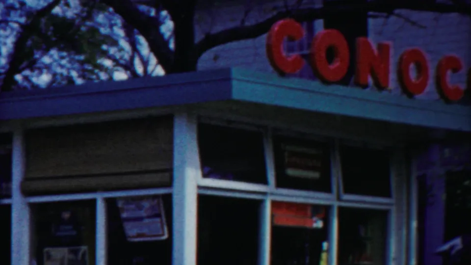 Angled 1958 view of the Conoco station corner, showing red exterior lettering, large glass panes, window placards, and the roof edge.