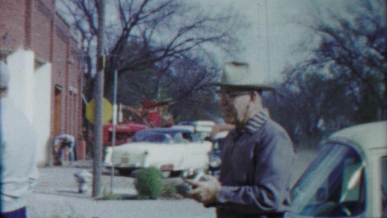 Two adult men stand near parked cars outside a brick building on a small-town street.