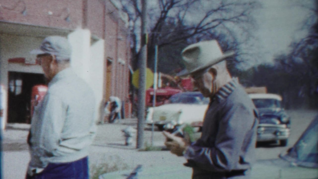 A man in a white cap walks past a man in a brimmed hat holding a small object.