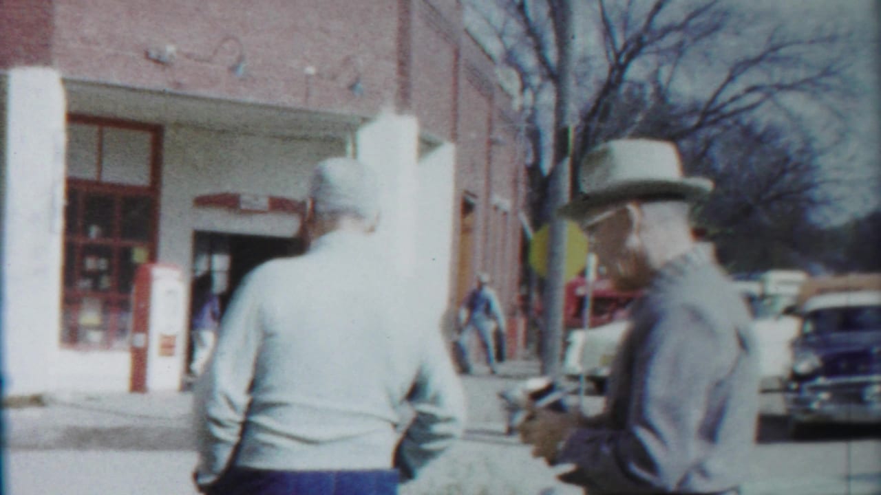 Two adult men remain on the sidewalk in Cheyenne County, Kansas, with a brick storefront, red fixture, and classic car behind them.