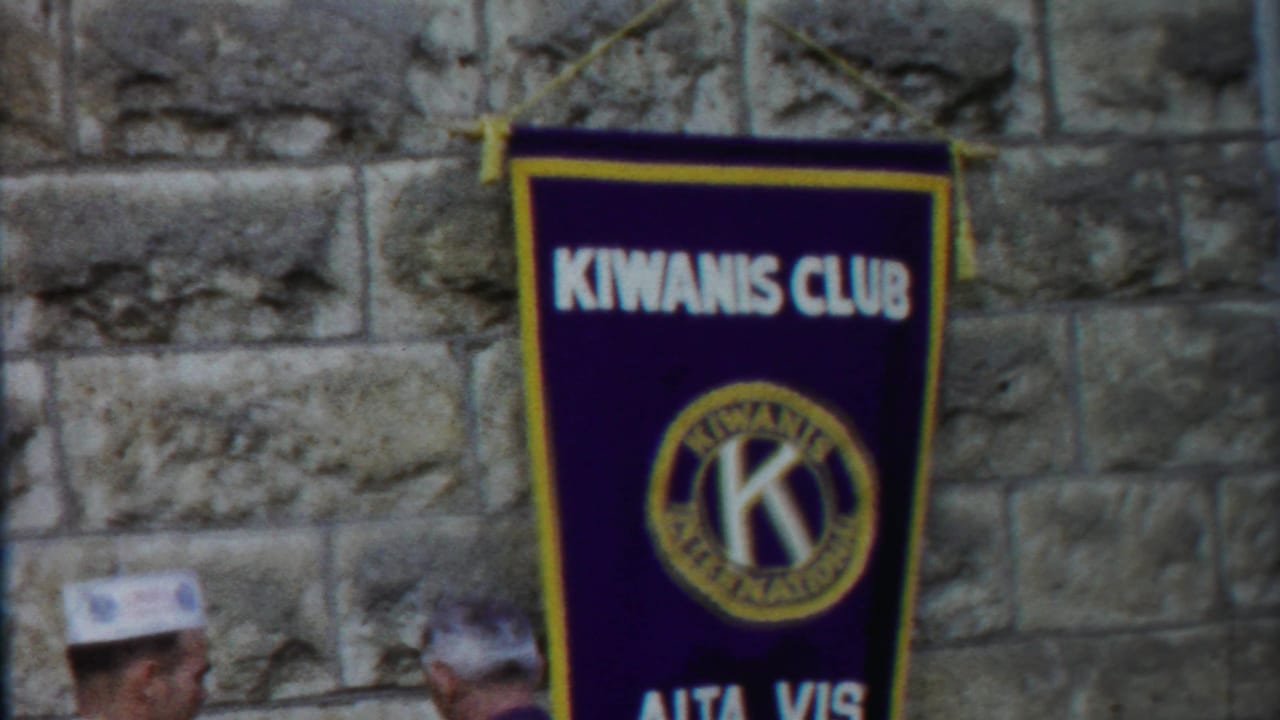 Purple Kiwanis Club banner on a stone wall with partial men at the lower edge.