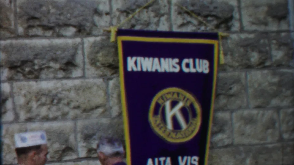 1958 Kansas home-movie frame showing a purple Kiwanis Club banner on a stone wall with two men partly visible below.