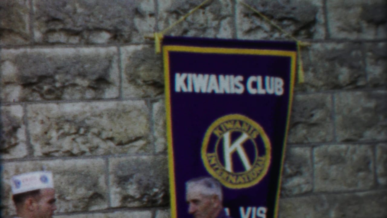 1958 Kansas frame with a Kiwanis Club banner centered on a stone wall and two men partly visible near the lower frame.