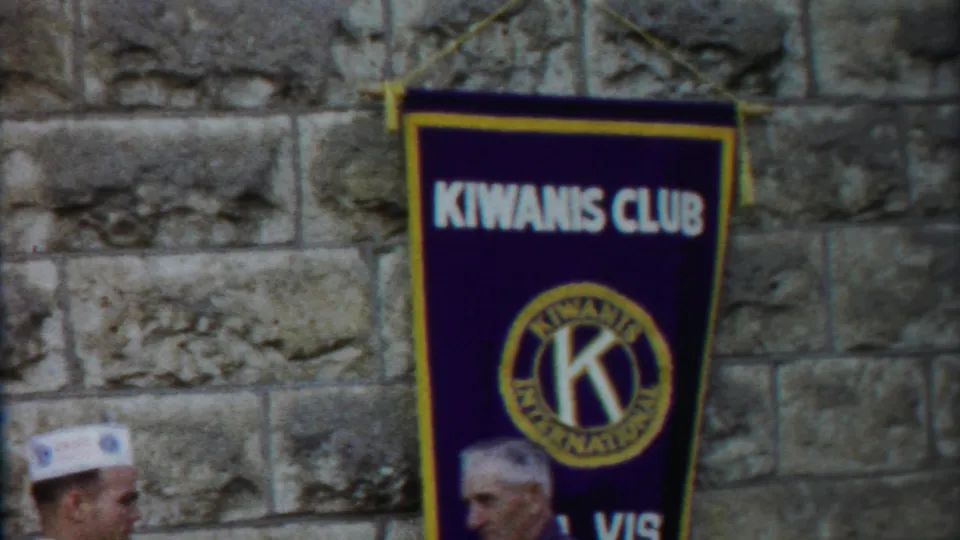 1958 Kansas frame with a Kiwanis Club banner centered on a stone wall and two men partly visible near the lower frame.