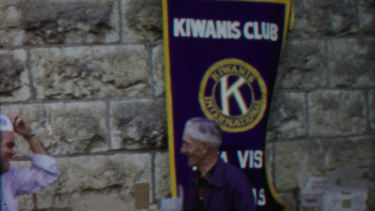 1958 Kansas home-movie frame showing a purple Kiwanis Club banner on a stone wall as older men stand nearby.