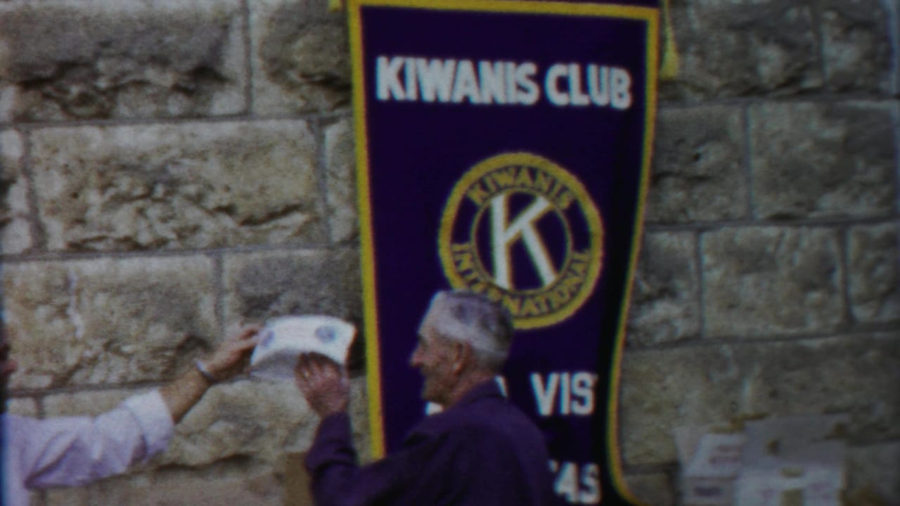 A man in a light shirt passes a paper to an older man near a Kiwanis Club banner in Cheyenne County, Kansas.