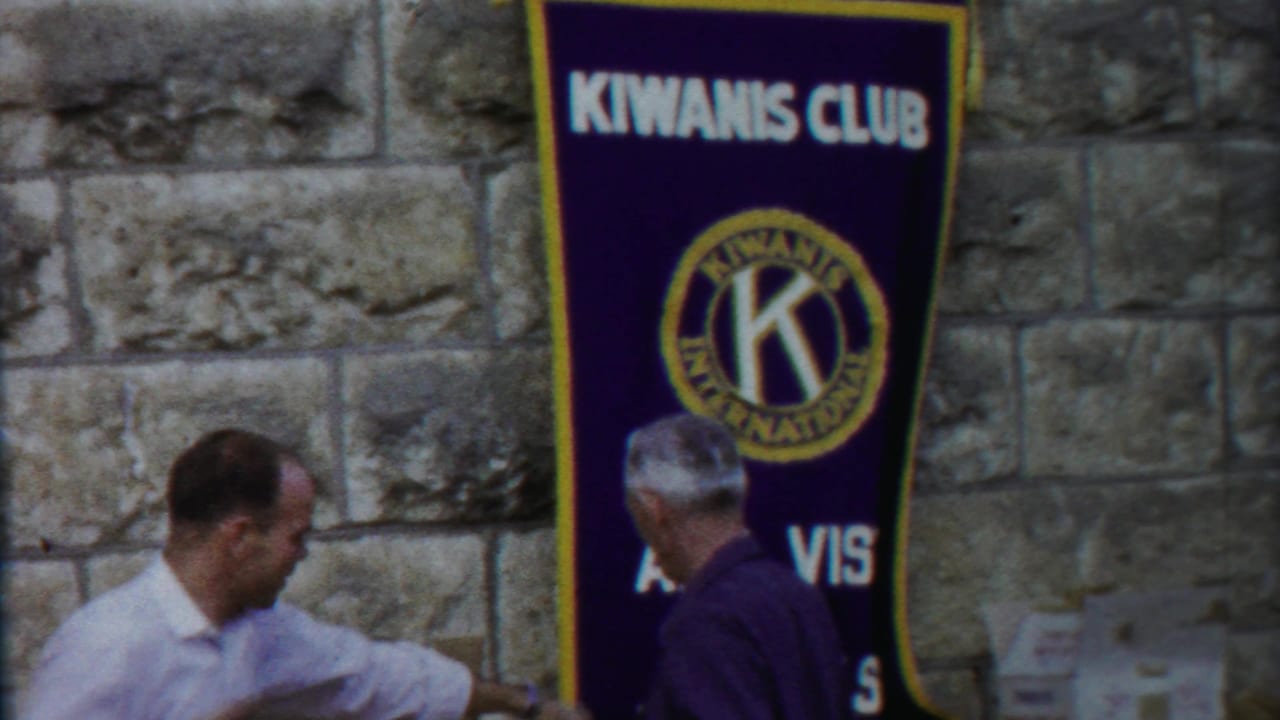 1950s 8mm frame of men near a large Kiwanis Club banner, with the stone wall and public-event setting visible.