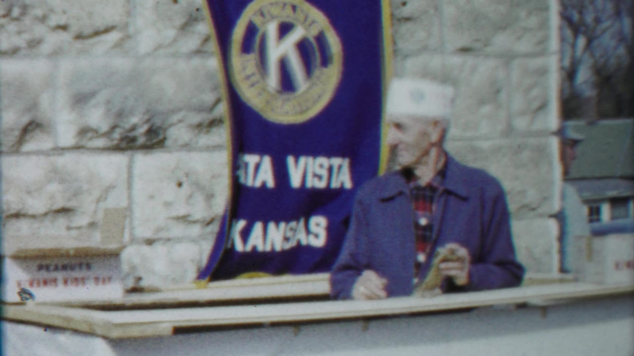 Older man in a white cap and blue jacket stands by a booth edge beside a Kiwanis banner in 1958 Kansas footage.