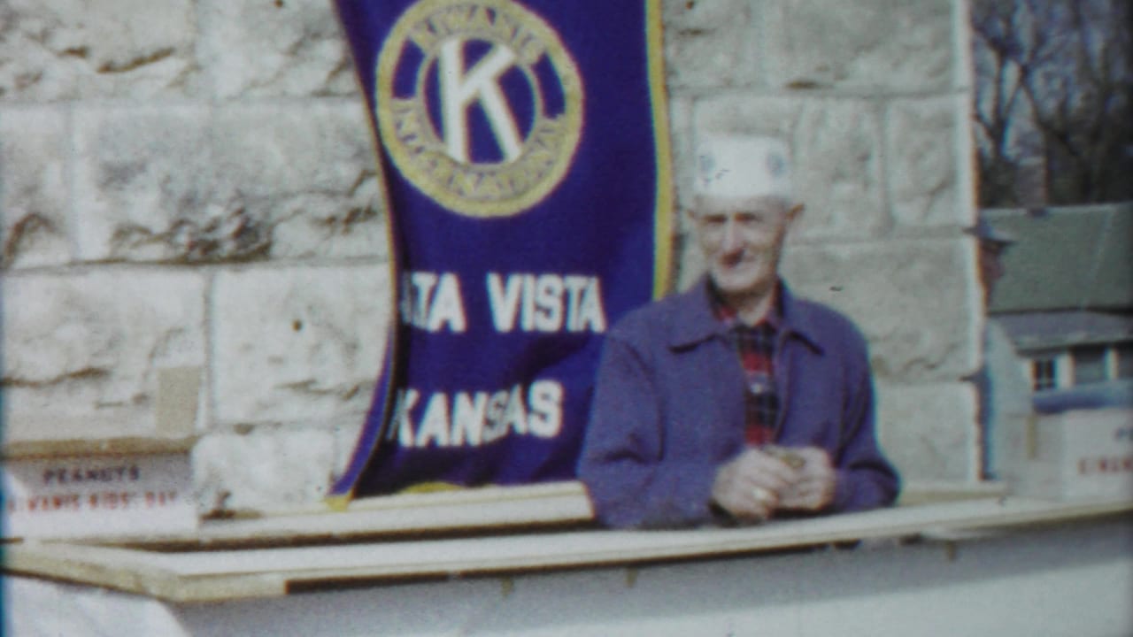 Older man in a white hat and blue jacket stands at a counter before an Alta Vista Kansas Kiwanis banner in 1958 home-movie footage.