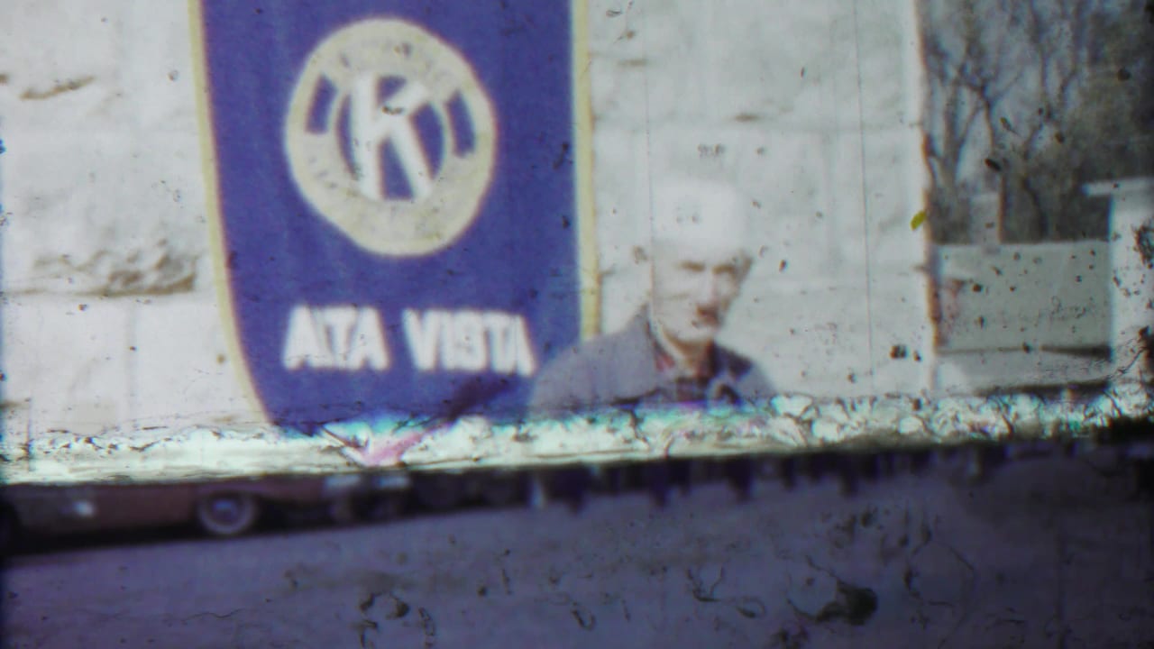 Alta Vista Kansas Kiwanis banner dominates the frame while the older man is partly visible beside the stone wall.
