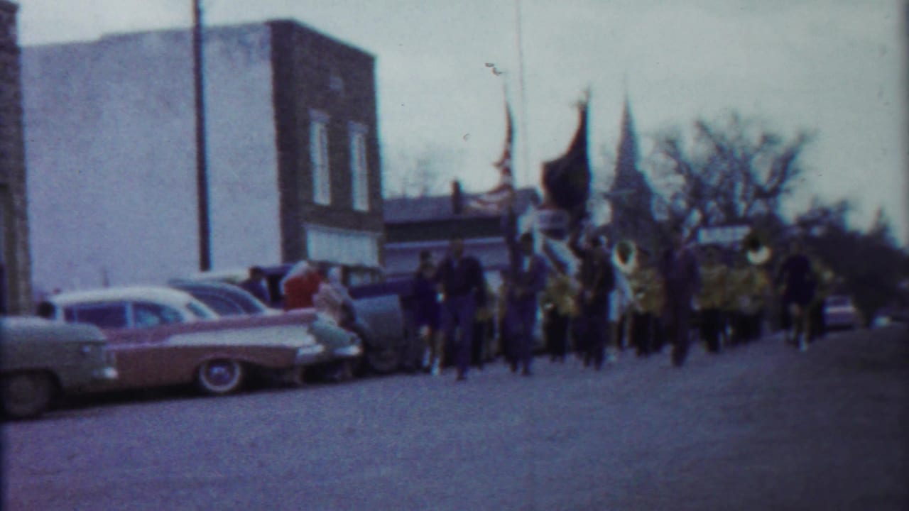 A 1958 Kansas parade view shows classic cars parked along the street as a marching band approaches in the distance.