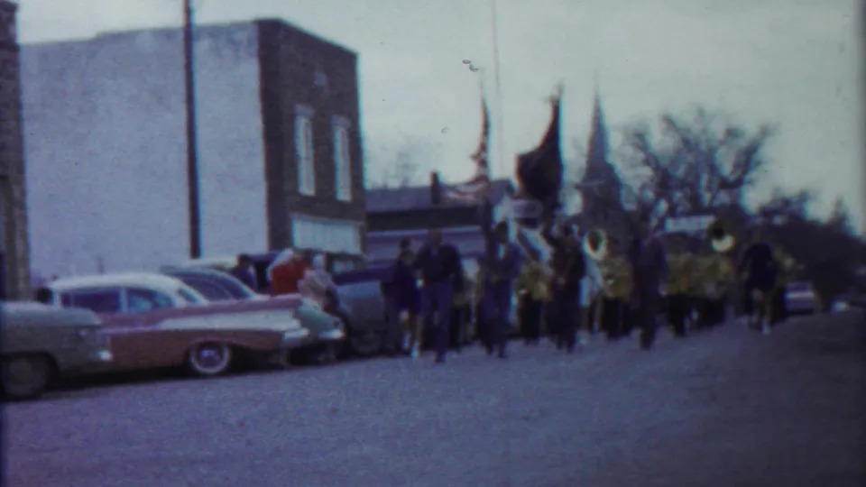 A 1958 Kansas parade view shows classic cars parked along the street as a marching band approaches in the distance.