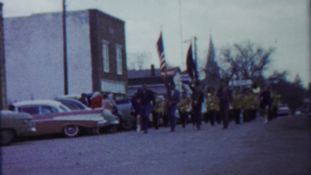 1958 Kansas home-movie frame showing a marching band and flags moving toward the camera past parked classic cars and brick buildings.