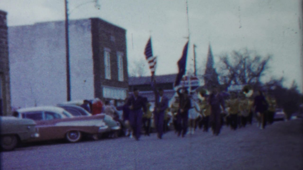 1958 Kansas parade footage frame with flag bearers, band members, spectators, and mid-century cars along a small-town street.