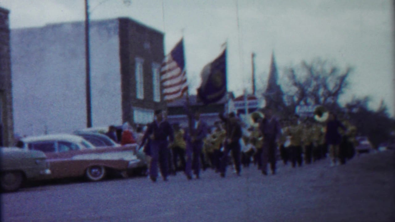 Closer 1958 Kansas parade frame with the American flag prominent, band members in uniform, brass instruments, and parked cars at left.