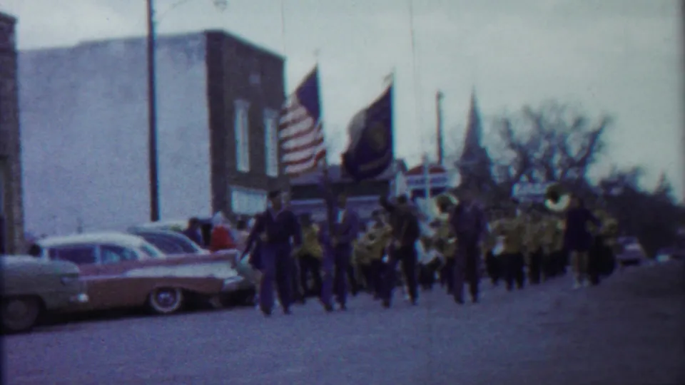 Closer 1958 Kansas parade frame with the American flag prominent, band members in uniform, brass instruments, and parked cars at left.