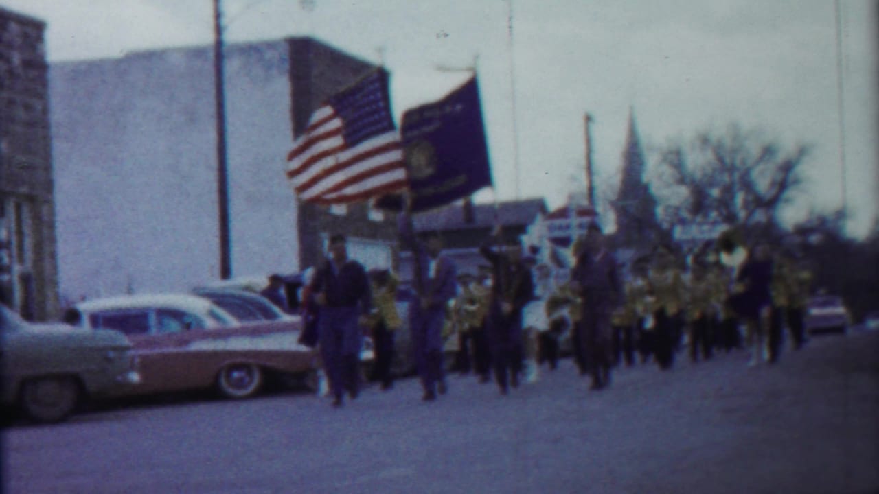 1958 Kansas home-movie frame showing parade flags, marching band members, classic cars, and a distant church steeple on a town street.