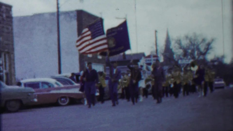 1958 Kansas home-movie frame showing parade flags, marching band members, classic cars, and a distant church steeple on a town street.