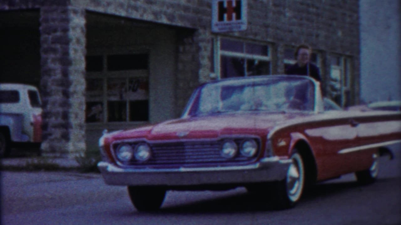 Red and white vintage convertible parked outside a stone building with a woman standing behind it.