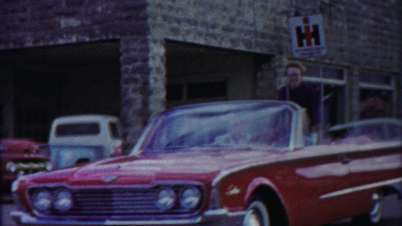 The red and white convertible moves through the frame near a stone building in Cheyenne County, Kansas, with a woman visible beside the car.