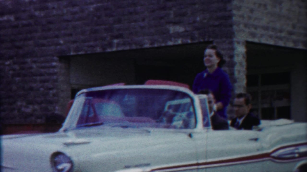 1958 Cheyenne County, Kansas home-movie frame showing a white convertible, woman in purple standing in back, and suited men riding past a stone building.