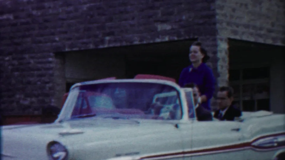 1958 Cheyenne County, Kansas home-movie frame showing a white convertible, woman in purple standing in back, and suited men riding past a stone building.