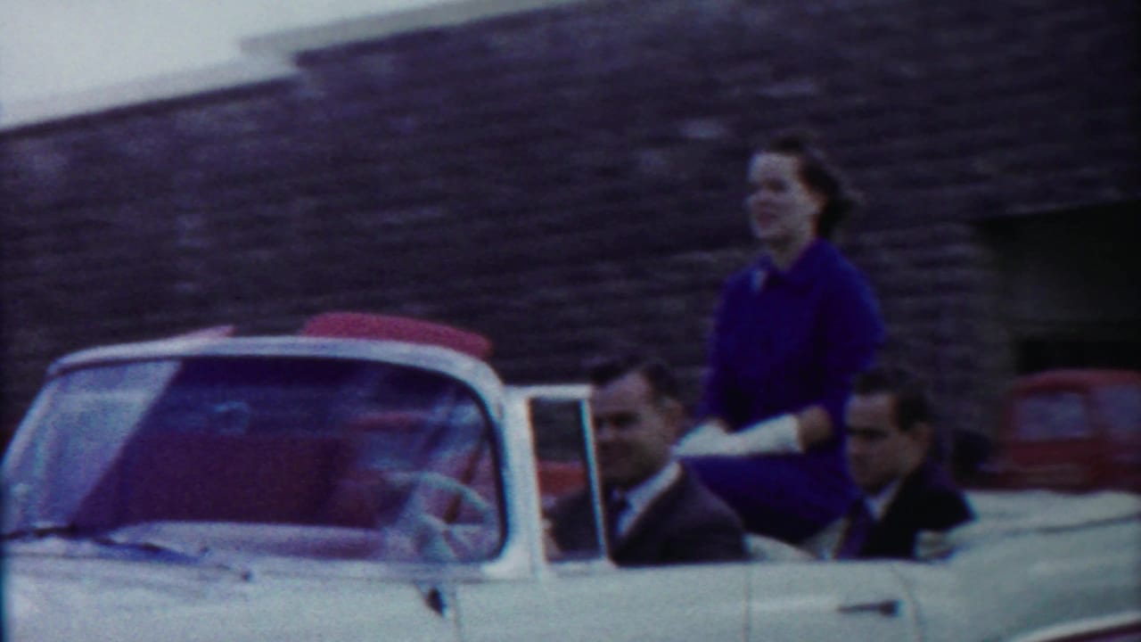 White convertible with red interior moving past a stone building as a woman stands in the rear seat.