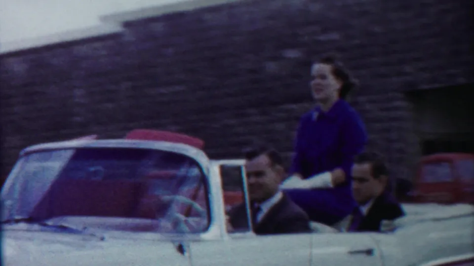 The convertible continues through the town street, with a standing woman in purple and seated men visible against the stone building backdrop.