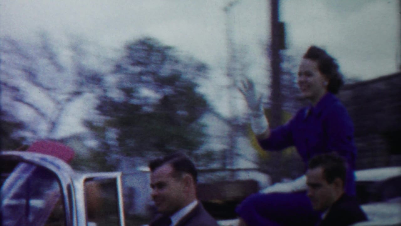 Home-movie frame from the moving street scene showing the woman smiling and waving from the open convertible as trees blur in the background.