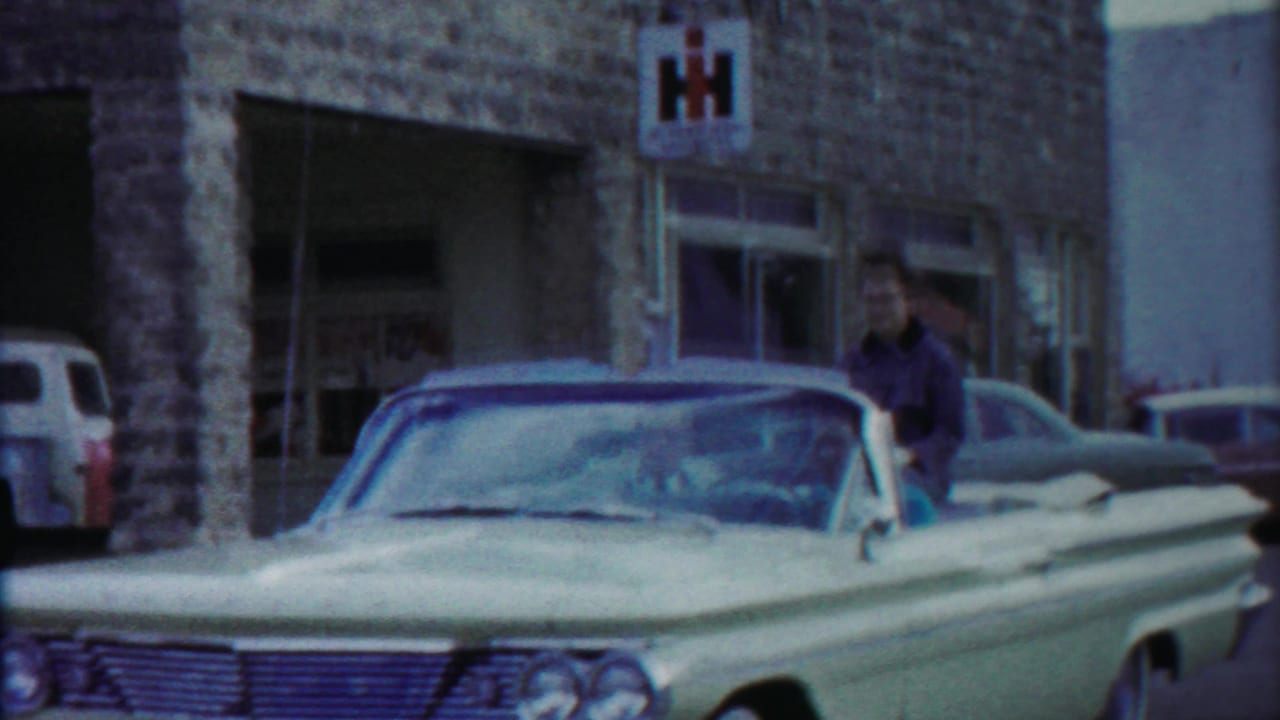 The car approaches or passes the camera near a stone-front building, with an IH sign, parked vehicles, and passengers visible in the convertible.