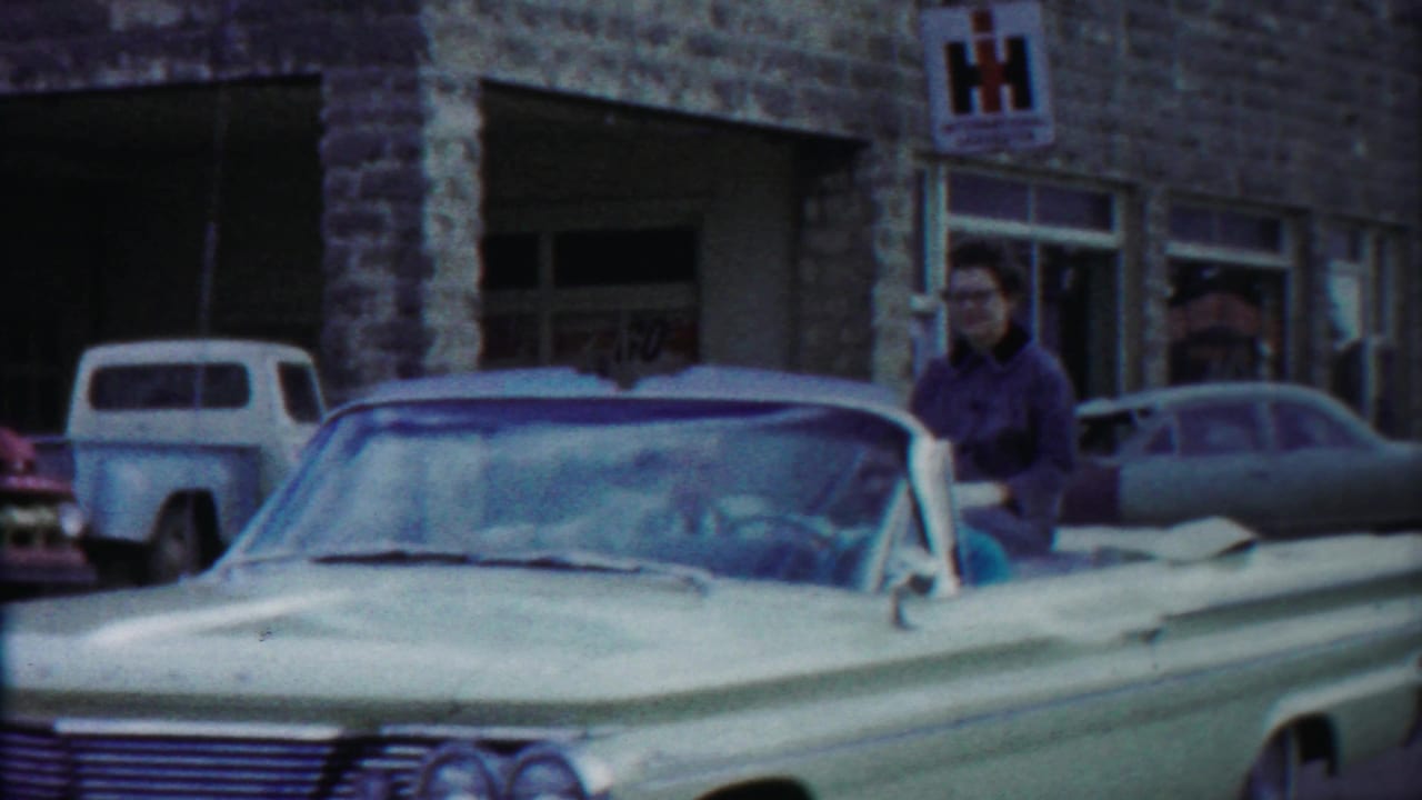 1958 Kansas home-movie frame showing a woman by a light convertible near a stone storefront with an International Harvester sign.