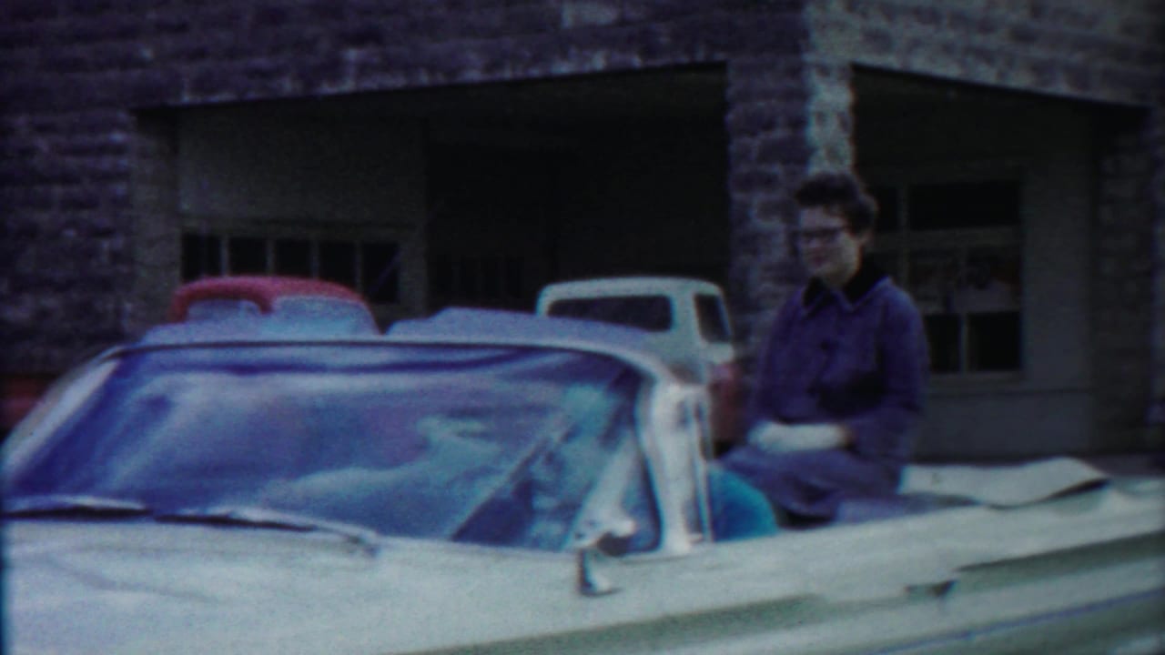 A woman in glasses sits in a light-colored convertible on a Cheyenne County street, with 1950s cars and a storefront behind her.