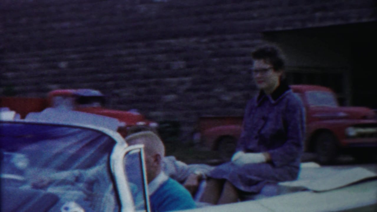 Home-movie frame from 1958 Kansas showing a woman in a convertible, a partial passenger, and parked vehicles outside a stone building.