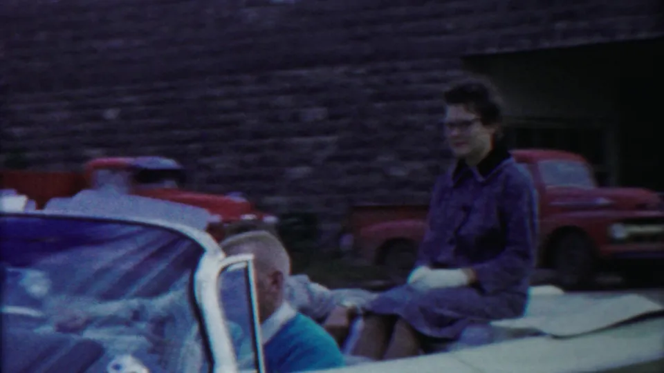 Home-movie frame from 1958 Kansas showing a woman in a convertible, a partial passenger, and parked vehicles outside a stone building.