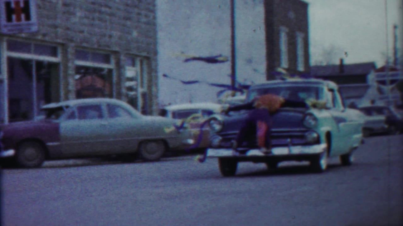 1958 Kansas street scene with an IH sign, parked mid-century cars, and a man leaning over the hood of a turquoise vehicle.